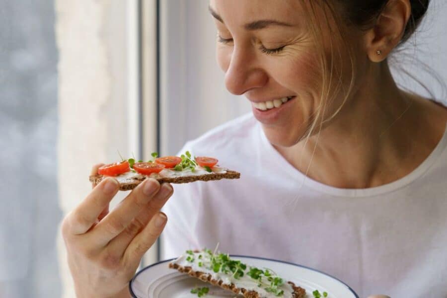Smiling woman eating rye crispbread with tofu cheesy spread, tomatoes and microgreens